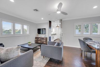 Bright open-plan living and dining room with gray sofas, black coffee table, rustic wood media cabinet, wall-mounted TV, hardwood floors, ceiling fan, and white plantation shutters overlooking greenery — modern coastal-style home interior.