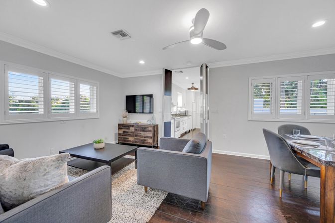 Bright open-plan living and dining room with gray sofas, black coffee table, rustic wood media cabinet, wall-mounted TV, hardwood floors, ceiling fan, and white plantation shutters overlooking greenery — modern coastal-style home interior.