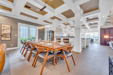 Open-concept waterfront dining room in a coastal-style home with a large wooden table set for eight, mid-century chairs, coffered ceiling, modern pendant light, and large windows overlooking a marina.