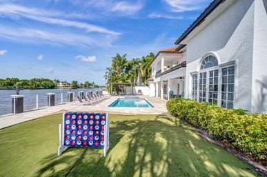Coastal luxury home with private pool, dock and sun loungers by a palm-lined waterfront, lawn with a giant outdoor disc-drop game under a blue sky.