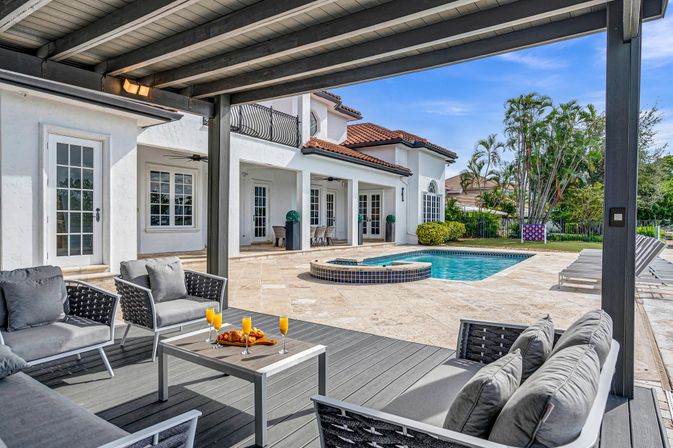Covered outdoor patio with gray lounge seating and a table of orange mimosas, overlooking a tiled pool and spa beside a two-story white Mediterranean-style home with red tile roof and palm trees