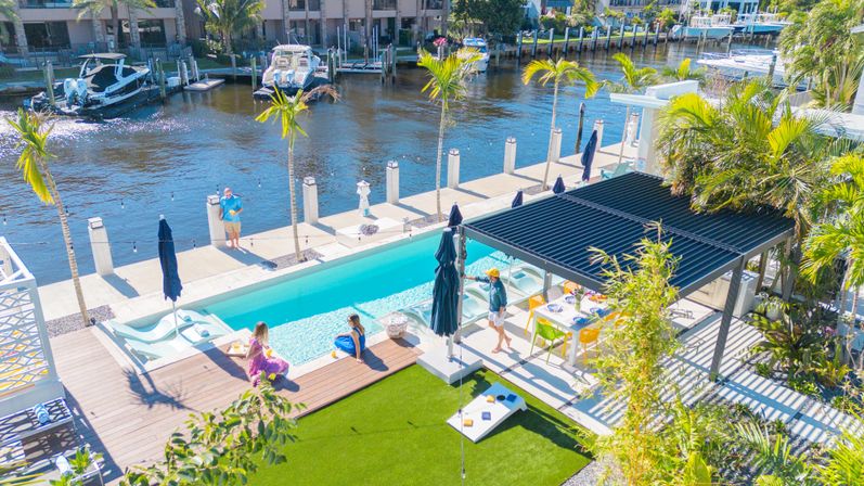Aerial view of a sunny canal-side waterfront backyard with a turquoise lap pool, wooden deck and loungers, palm trees, pergola-covered outdoor dining, docked boats and people relaxing by the water.