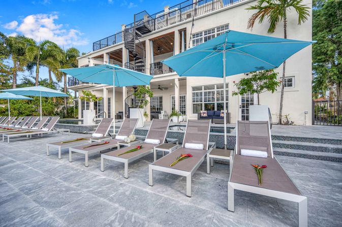Sun-ready poolside lounge chairs with rolled towels and red tropical flowers under bright blue umbrellas on a stone patio in front of a multi-level coastal villa surrounded by palm trees