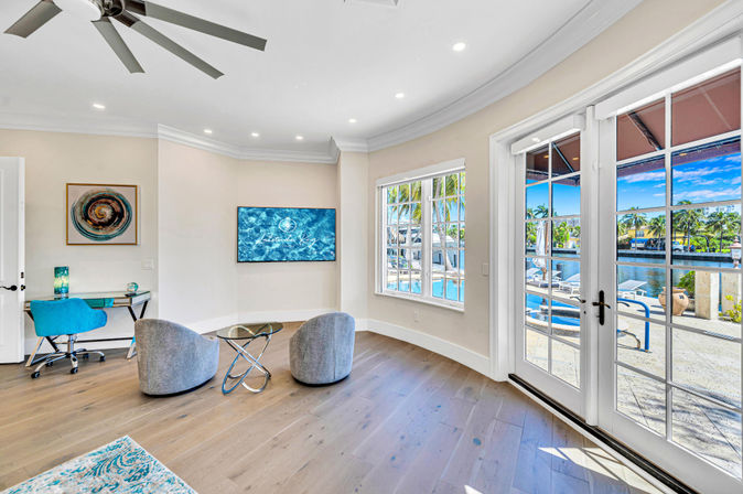Bright modern coastal living room with hardwood floors, two gray lounge chairs, glass coffee table, wall-mounted TV and turquoise desk; French doors open to a sunny pool deck and palm-lined canal waterfront.