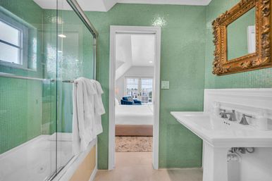 Bright green mosaic-tiled bathroom with glass-enclosed tub-shower and folded white towels, white pedestal sink beneath an ornate gold mirror, opening to an ensuite sunlit bedroom with white bedding and a blue accent chair.