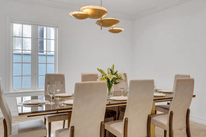 Sunlit contemporary dining room set for six with glass-top table, beige upholstered chairs, gold disc chandelier and a lily centerpiece.