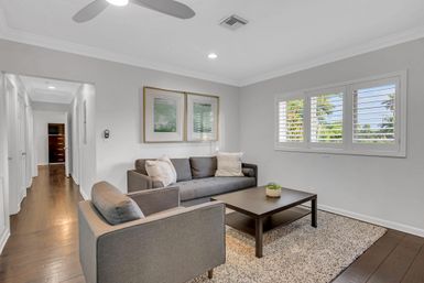 Cozy modern coastal living room with gray sofa and armchair, dark wood coffee table on a textured rug, hardwood floors and white plantation shutters framing palm trees outside.