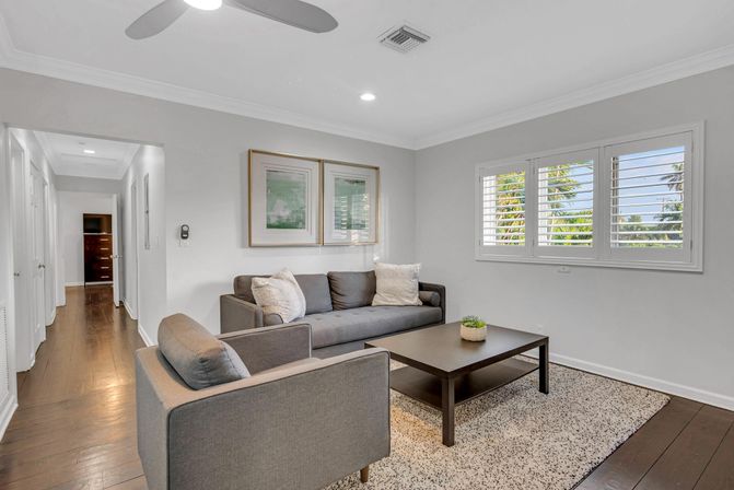Cozy modern coastal living room with gray sofa and armchair, dark wood coffee table on a textured rug, hardwood floors and white plantation shutters framing palm trees outside.