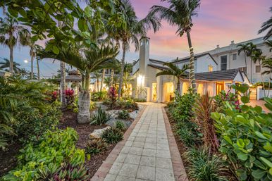 Sunset-lit tropical villa exterior with palm-lined paved walkway, lush landscaped garden, and warm Mediterranean-style architecture