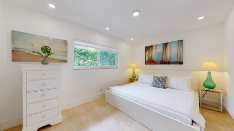 Bright modern coastal bedroom with white walls and tile floor, low white platform bed, green glass bedside lamps, beach artwork and a leafy window