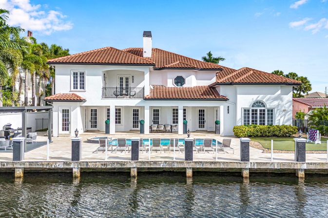 Sunny canal-front Mediterranean-style two-story villa with terracotta tile roof, balcony, palm trees, poolside lounge chairs, spacious patio and private dock on calm water.