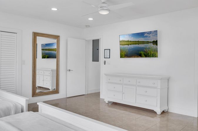 Bright white coastal-style bedroom with twin beds, large wood-framed mirror, wall-mounted TV showing a lake scene, white dresser on glossy tile floor, and ceiling fan.