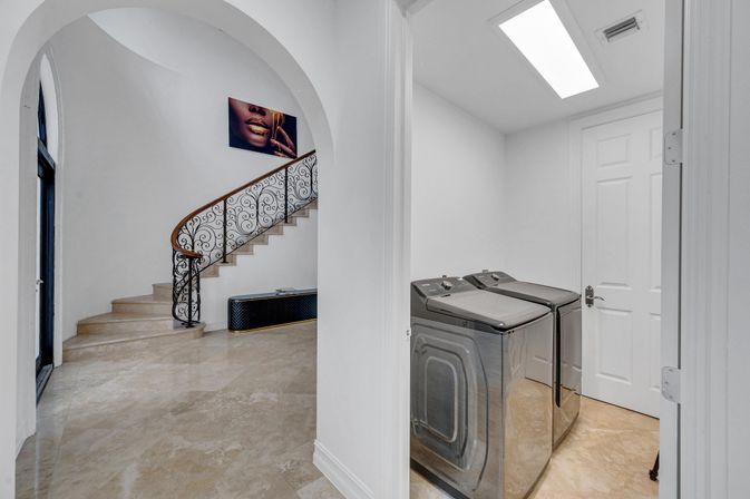 Elegant home foyer with curved marble staircase and ornate wrought-iron railing, bold wall art of gold-accented lips, and an adjacent bright laundry room with stainless steel washer and dryer under a skylight.