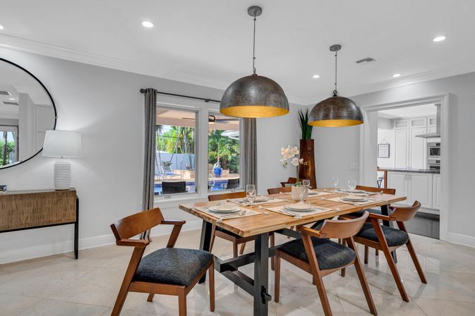 Bright, airy contemporary dining room with a mid-century wooden table set for six, angled wooden chairs, two oversized metal pendant lights, and a window view of a backyard pool and patio.