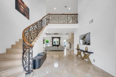 Sunlit luxury foyer with curved marble staircase and ornate wrought-iron railing, glossy stone floors, sleek console table, and open living room with a stone fireplace visible beyond.