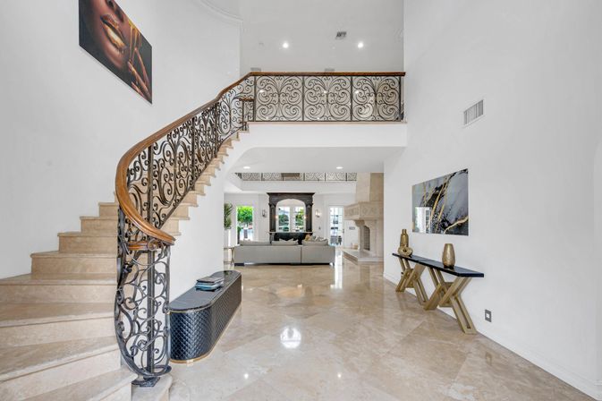 Sunlit luxury foyer with curved marble staircase and ornate wrought-iron railing, glossy stone floors, sleek console table, and open living room with a stone fireplace visible beyond.