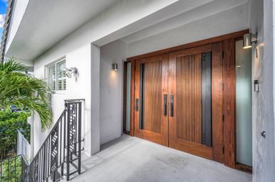 Recessed entry of a modern coastal home with warm wooden double doors, white stucco walls, black metal railing, palm fronds and wall sconces on a concrete porch.