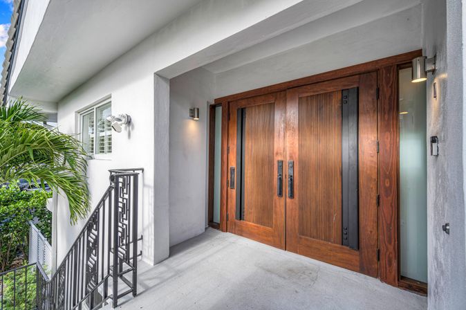 Recessed entry of a modern coastal home with warm wooden double doors, white stucco walls, black metal railing, palm fronds and wall sconces on a concrete porch.