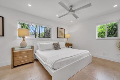 Bright, airy modern bedroom with white platform king bed, crisp linens, wooden nightstands with lamps, ceiling fan, tiled floor, and windows revealing lush tropical greenery outside.