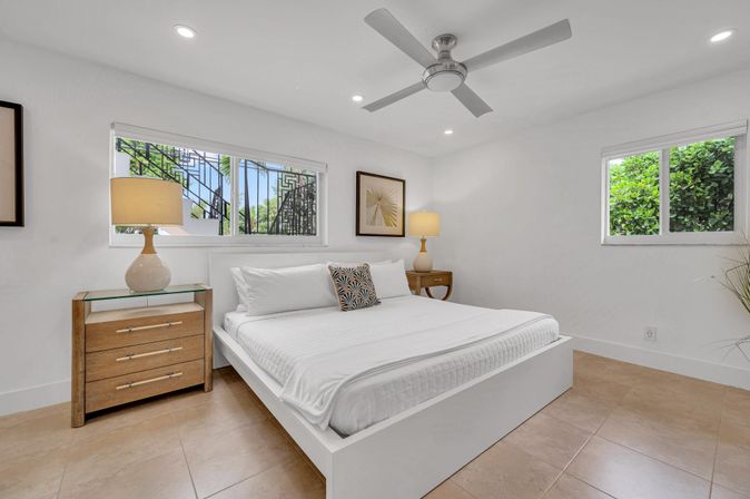 Bright, airy modern bedroom with white platform king bed, crisp linens, wooden nightstands with lamps, ceiling fan, tiled floor, and windows revealing lush tropical greenery outside.