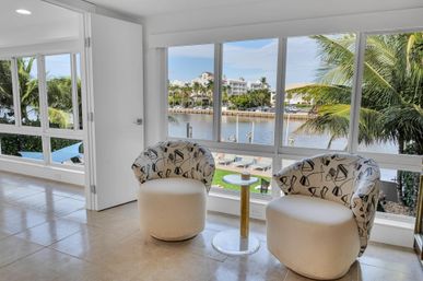 Cozy coastal sitting area in a bright waterfront condo: two patterned swivel chairs and a glass-top side table by floor-to-ceiling windows overlooking a palm-lined marina and waterfront residences.