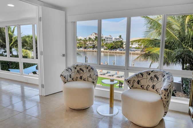 Cozy coastal sitting area in a bright waterfront condo: two patterned swivel chairs and a glass-top side table by floor-to-ceiling windows overlooking a palm-lined marina and waterfront residences.