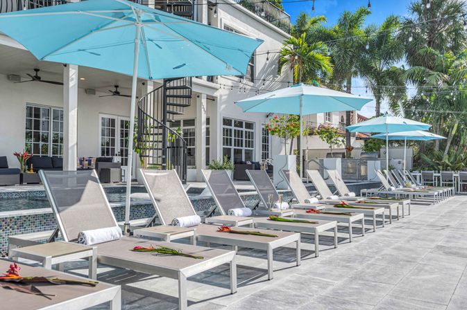 Tropical resort-style poolside with turquoise umbrellas shading a row of gray sun loungers topped with rolled towels and flowers, modern white villa with spiral staircase, palm trees and string lights overhead.