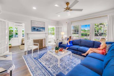 Sunlit coastal-style living room with large windows, oversized blue L-shaped sectional, patterned blue area rug, white coffee table, fireplace, ceiling fan, and two people relaxing.