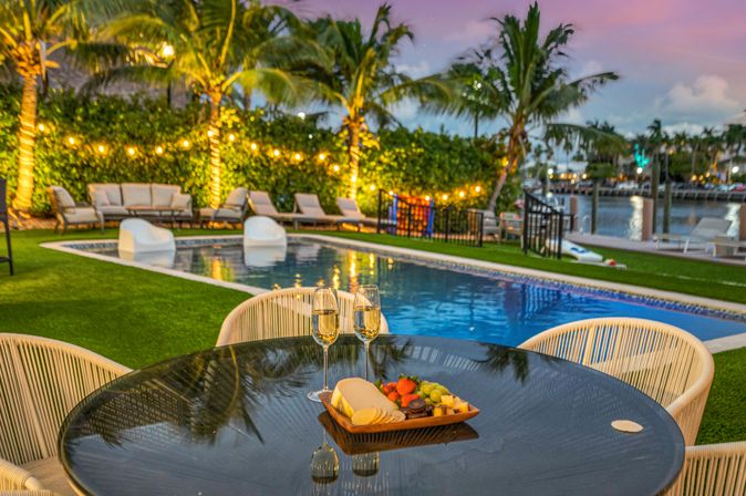 Poolside tropical waterfront scene at sunset with string-lit palm trees and lounge chairs, glossy table in foreground holding two champagne flutes and a cheese-and-fruit platter.