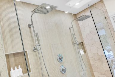 Glass-enclosed spa-like shower with dual chrome rain showerheads, vertical beige tile, hexagonal accent wall and recessed niches with bottles