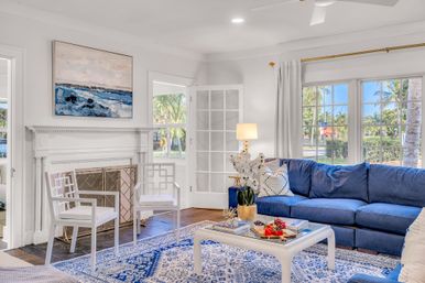 Sunlit coastal-style living room with a blue sectional, white fireplace, patterned blue rug, white armchairs and a coffee table with fruit; large windows and French doors reveal palm trees and a tropical street view.