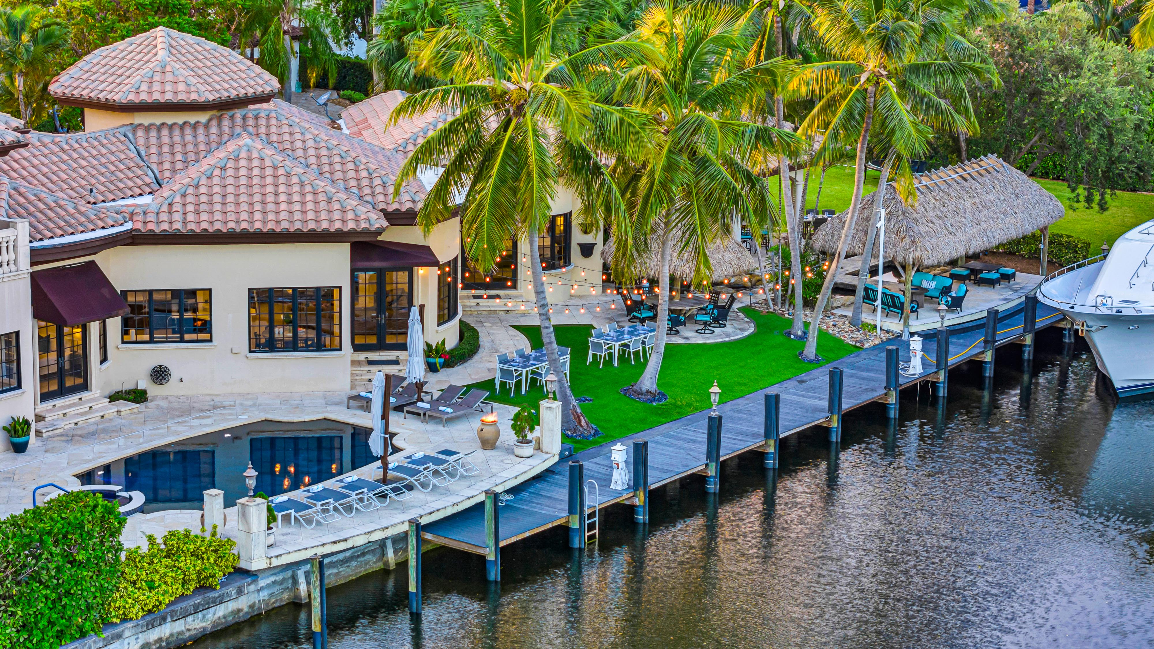 Waterfront tropical villa with terracotta roof, pool terrace, palm trees and tiki hut overlooking a wooden dock and moored yacht, string lights and outdoor seating