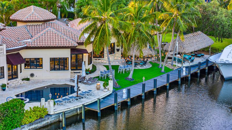 Waterfront tropical villa with terracotta roof, pool terrace, palm trees and tiki hut overlooking a wooden dock and moored yacht, string lights and outdoor seating