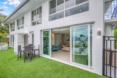 Modern coastal waterfront patio with artificial turf, black wicker bistro set and sliding glass doors opening to a bright living room and canal view with palm trees.