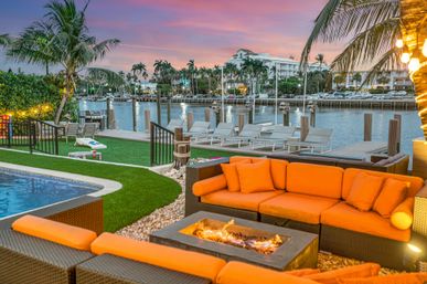 Waterfront tropical patio at sunset with bright orange outdoor sofa and fire pit, pool and grassy yard leading to a dock and palm-lined marina