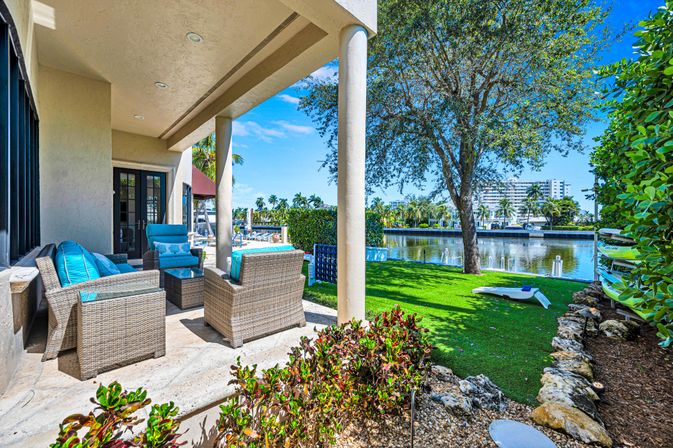 Sunny tropical waterfront patio with wicker seating and bright turquoise cushions overlooking a palm-lined canal, green lawn, tree and stacked kayaks.