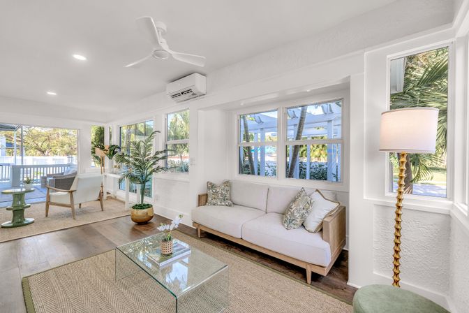 Bright coastal sunroom-style living room with white sofa, glass coffee table, tropical plants, wood floors and large windows overlooking a sunny patio