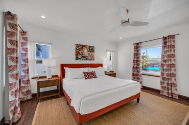Bright tropical vacation rental bedroom with king bed featuring an orange upholstered frame and white linens, patterned coral curtains, wooden nightstands with lamps, ceiling fan and a window showing a sunny pool and palm tree view.