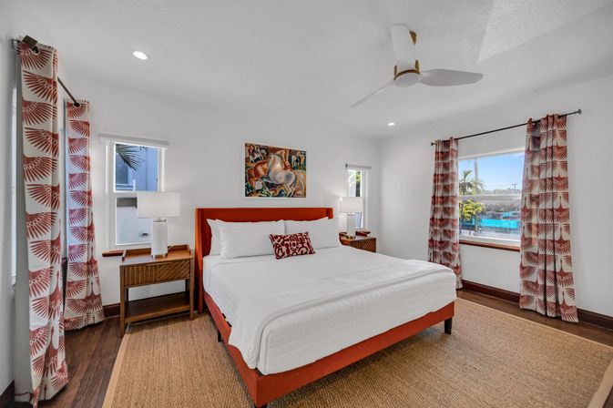 Bright tropical vacation rental bedroom with king bed featuring an orange upholstered frame and white linens, patterned coral curtains, wooden nightstands with lamps, ceiling fan and a window showing a sunny pool and palm tree view.
