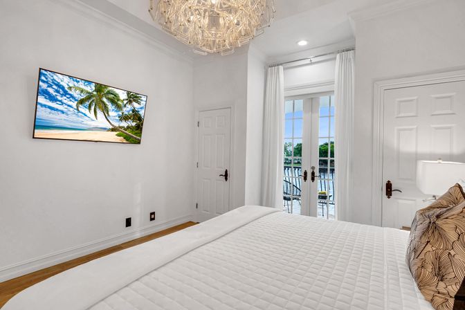 Bright white waterfront bedroom with quilted bed and patterned pillow, wall-mounted TV showing a tropical beach, crystal chandelier, and French doors opening to a balcony with outdoor seating and water views.
