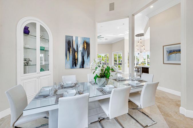Sunlit open-concept dining room in a luxury home with a glass table set for six, sleek white chairs, a potted orchid centerpiece, abstract blue-and-gold wall art, built-in glass-front cabinet, and views into a bright kitchen and outdoor area.