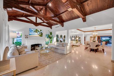 Sunlit luxury tropical villa living and dining space with dark exposed wooden beams and vaulted ceiling, polished marble floors, neutral sofas around a white fireplace, glass-and-gold coffee table, arched windows, lush indoor plants, and a gold chandelier over the dining area.