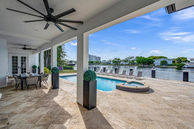 Waterfront pool patio with covered dining lanai, oversized ceiling fans, tile decking, built-in spa and a row of lounge chairs overlooking a calm canal and palm-lined shoreline.