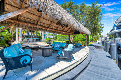 Sunny waterfront tiki-hut lounge with turquoise cushioned wicker sofas around a circular fire pit, shaded by a thatched palapa beside a wooden dock and moored yacht in a tropical marina.