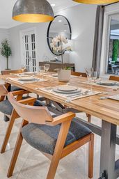 Modern dining room interior with a long wooden table set for six, mid-century wood chairs with gray cushions, an orchid centerpiece, plates and wine glasses, large dome pendant lights, round wall mirror and marble floor