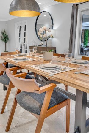 Modern dining room interior with a long wooden table set for six, mid-century wood chairs with gray cushions, an orchid centerpiece, plates and wine glasses, large dome pendant lights, round wall mirror and marble floor