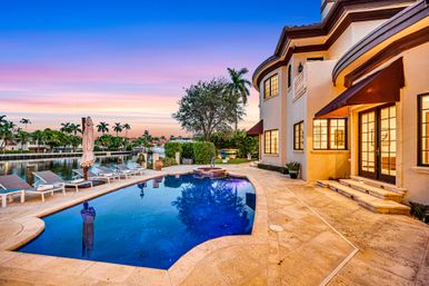 Sunset at a luxury waterfront home with a curved blue pool and spa, stone patio with loungers, palm-lined canal and docked boat