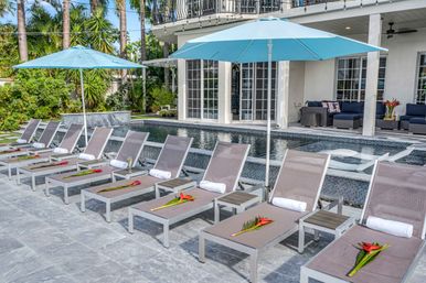 Row of modern sun loungers with rolled white towels and red tropical flowers under aqua umbrellas beside a mosaic-tiled pool on a tropical villa patio with palm trees