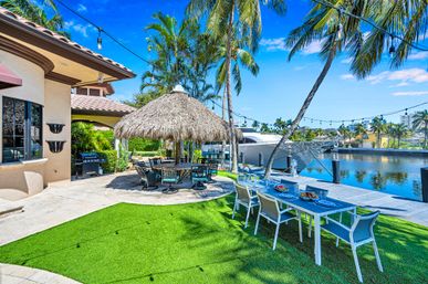 Sunlit waterfront tropical backyard with a thatched palapa dining area, outdoor dining table on green lawn, swaying palm trees, string lights and a docked yacht