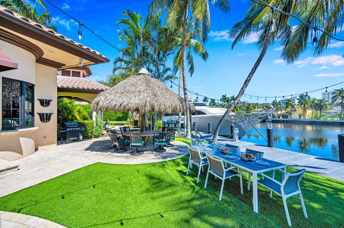 Sunlit waterfront tropical backyard with a thatched palapa dining area, outdoor dining table on green lawn, swaying palm trees, string lights and a docked yacht